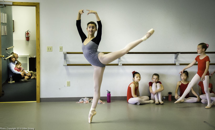 Studio Multiple Layers: Malerie Moore in a pique arabesque while behind her are parents in the hallway and younger dancers both sitting along the edges and going on.